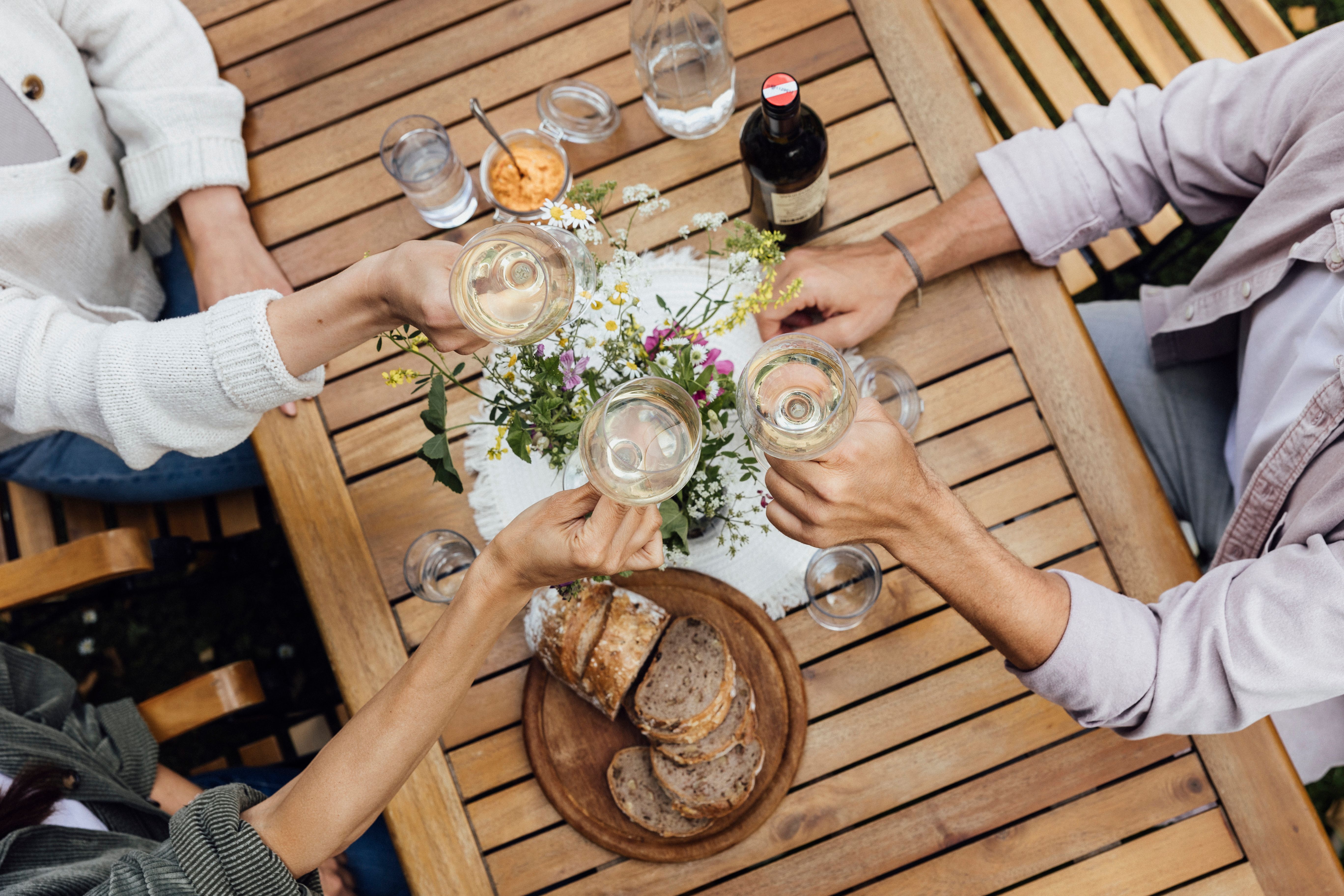 Three people clink glasses of wine at a wooden table with bread and flowers on it.