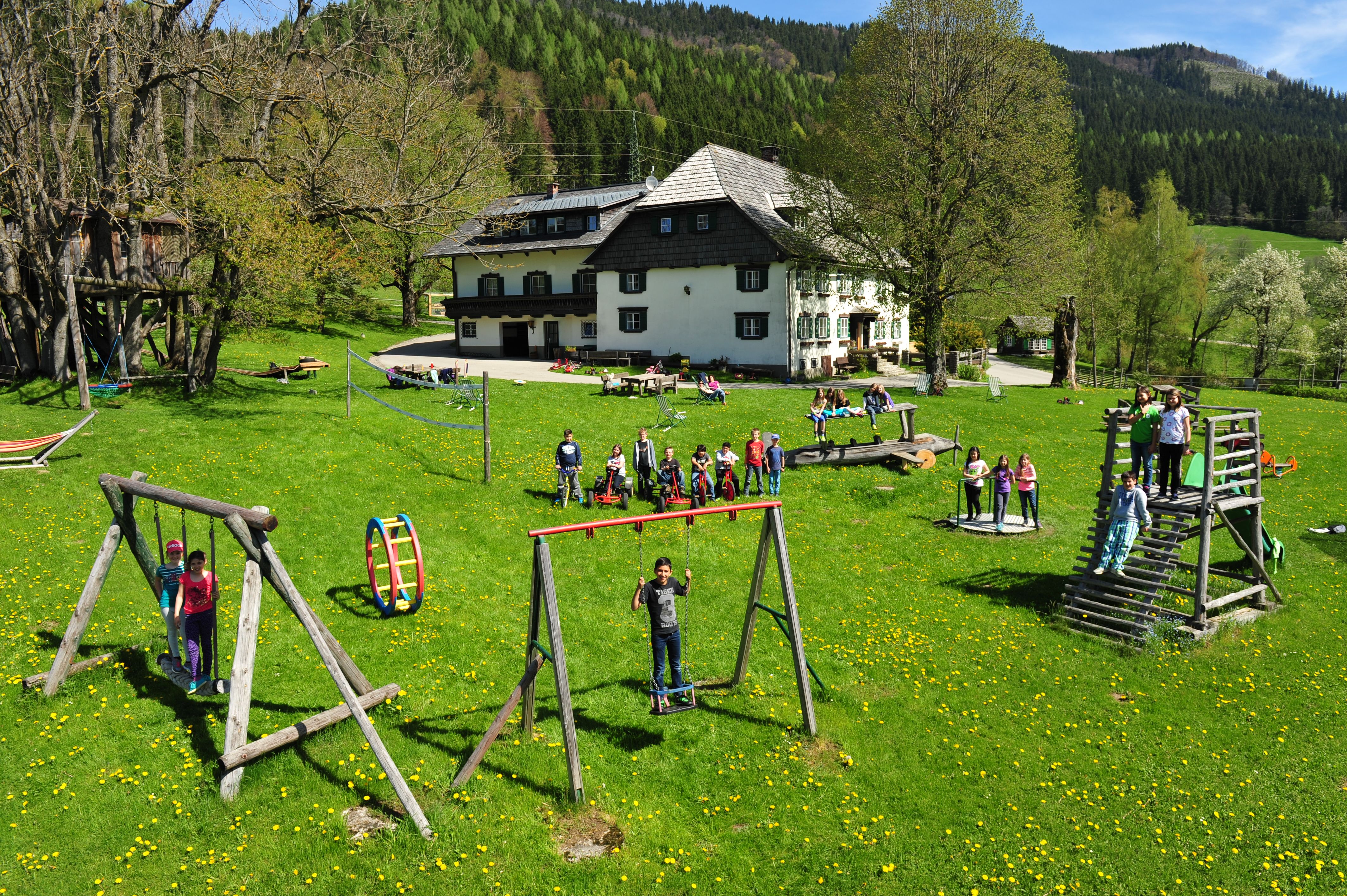 Children play on a playground in front of a large house in a green landscape.