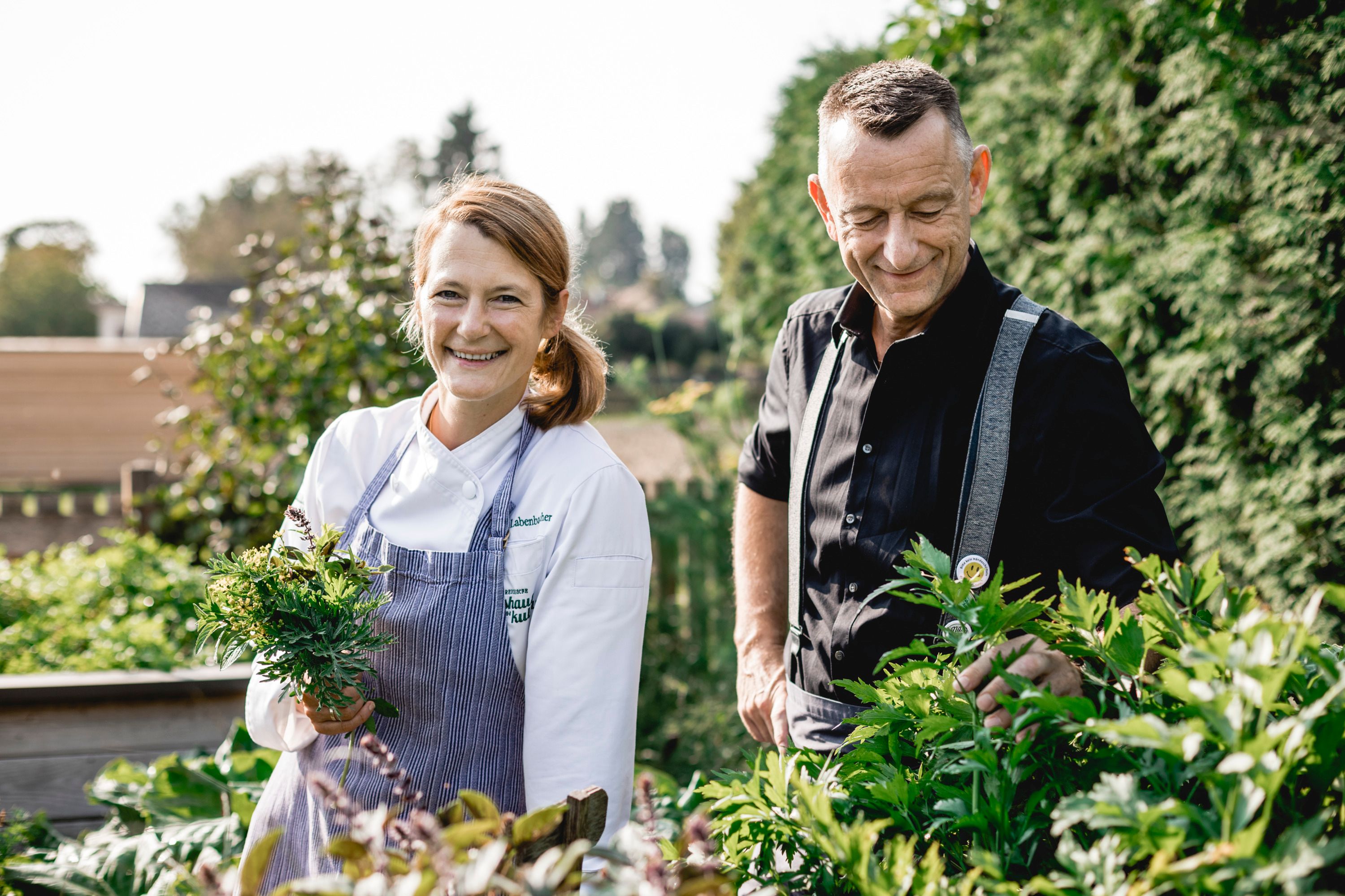 Two people are standing in a garden, a woman in cooking clothes is holding herbs.