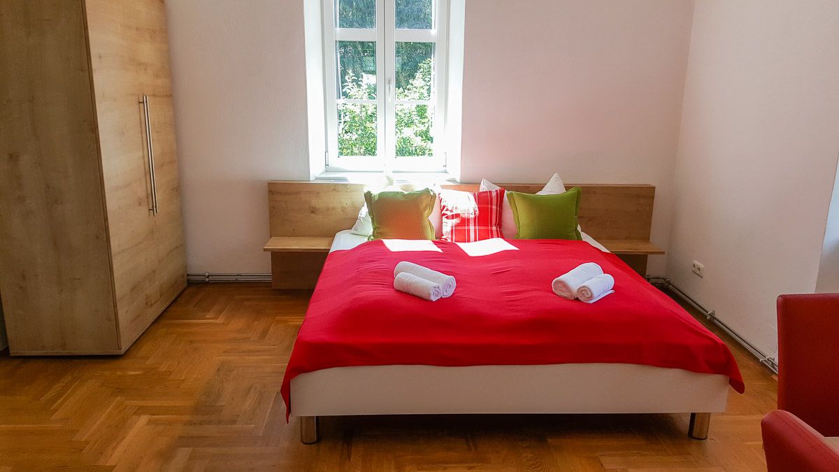 Bedroom with red bedspread, pillows and towels on the bed, wooden wardrobe and window in the background.