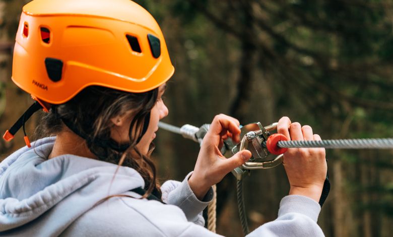 Person wearing an orange helmet attaches safety equipment to a rope in the forest ropes course.