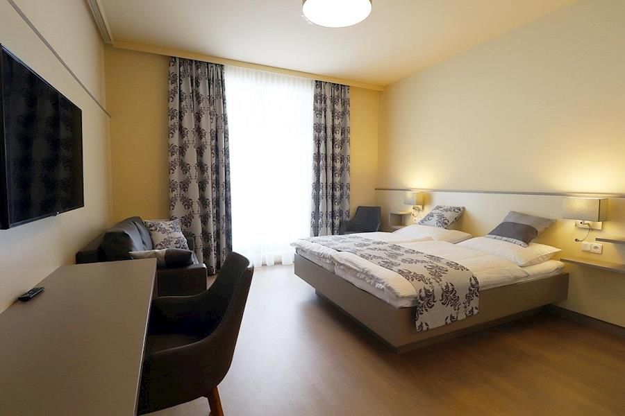 Modernly furnished hotel room with double bed, desk, TV and armchairs.