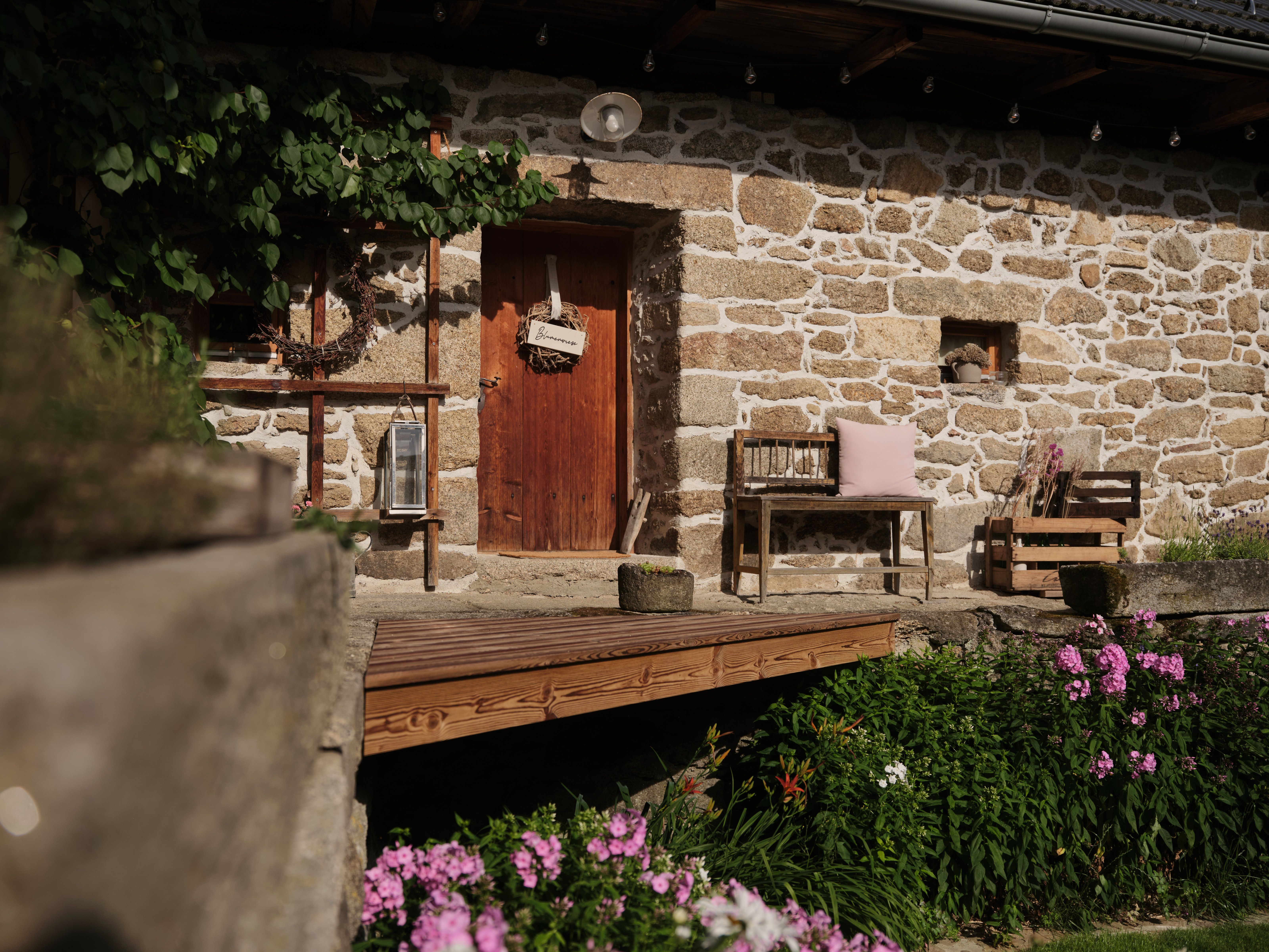 Rustic stone house with wooden door, bench and flowers in the foreground.