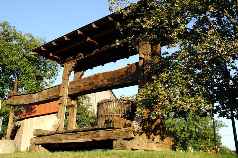 Old wooden wine press outdoors, surrounded by trees and buildings.