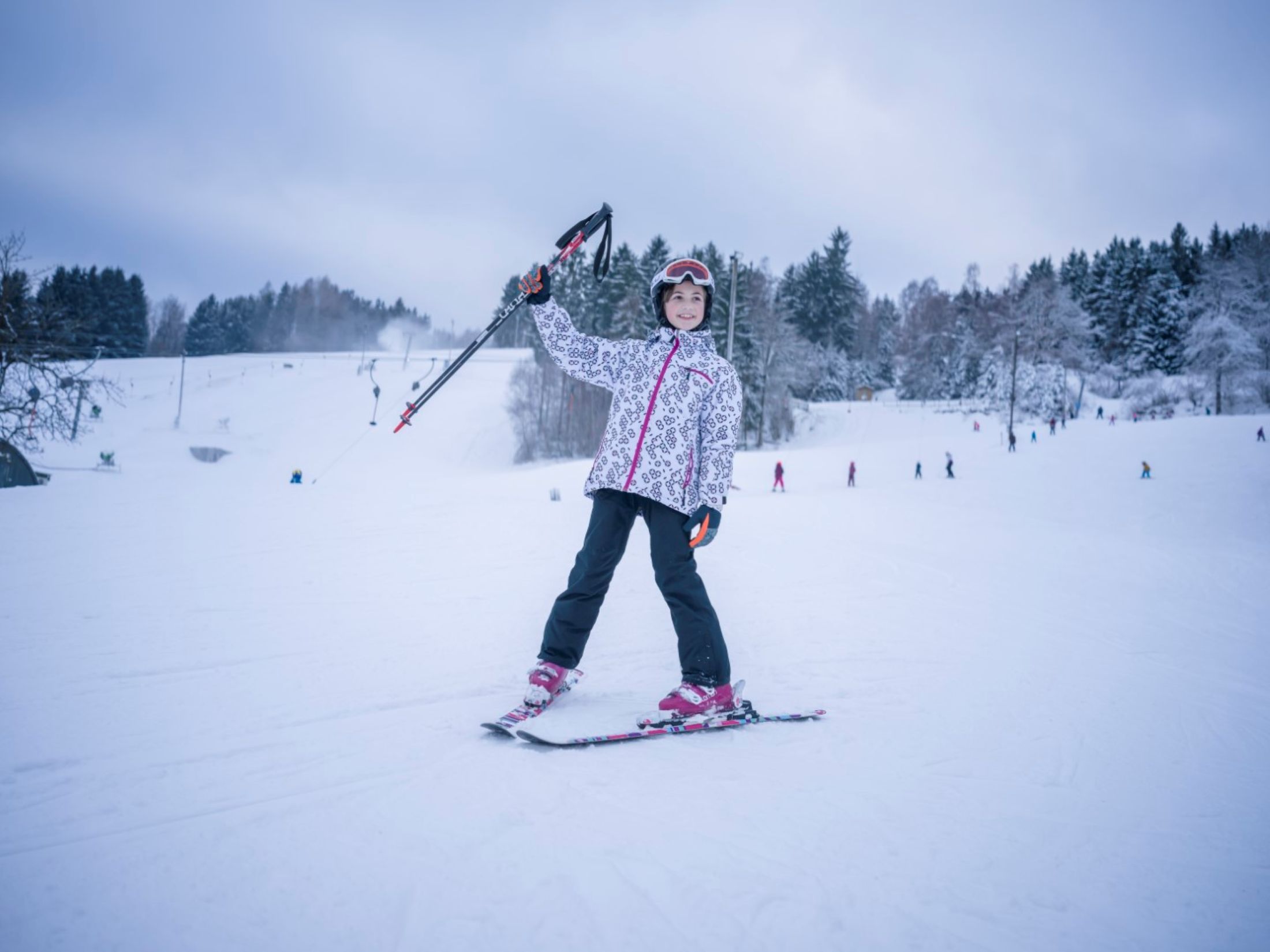 A child in ski equipment stands on a snowy slope and holds ski poles in the air. Trees and other skiers can be seen in the background.
