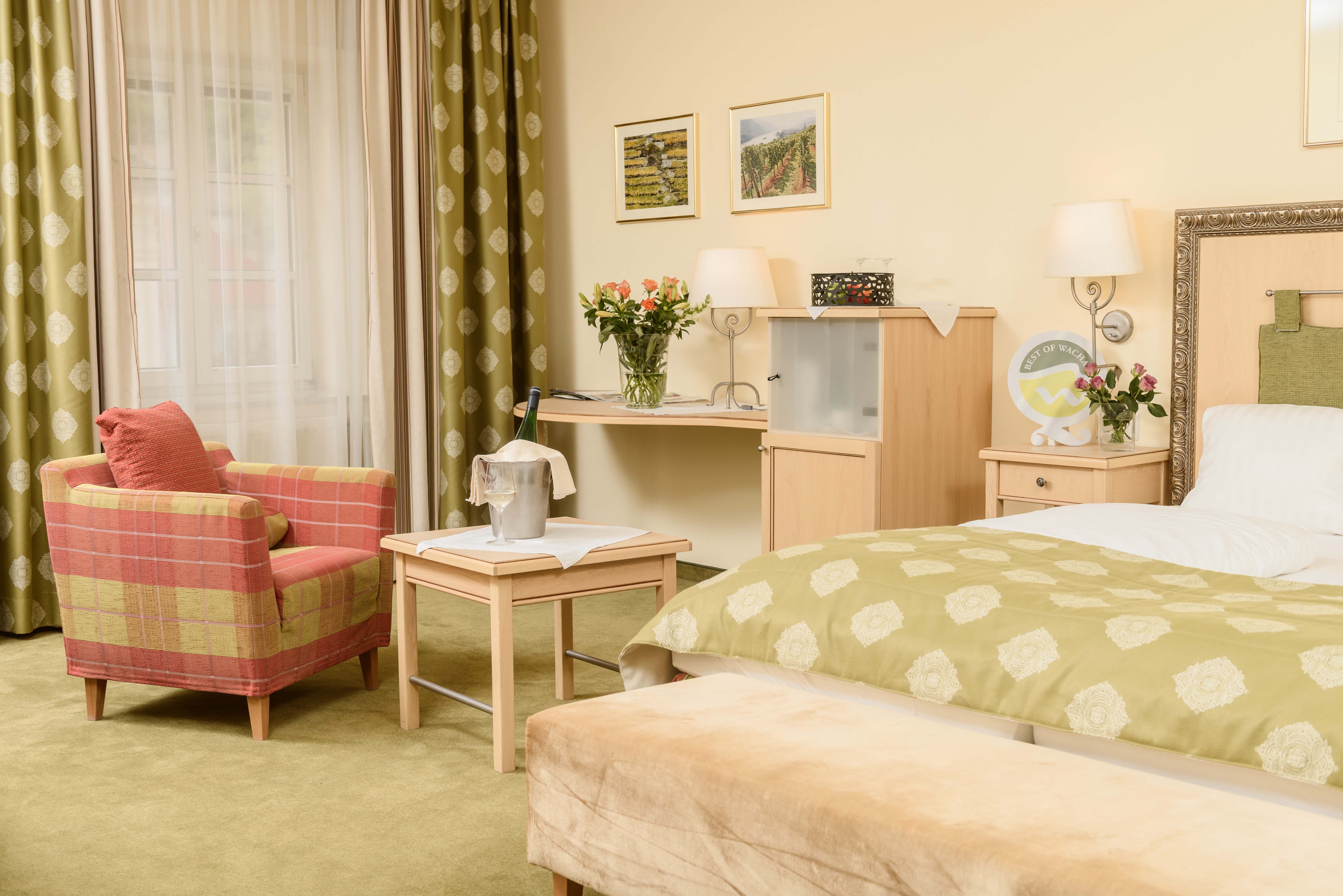 Cozy hotel room with green carpet, red armchair, table with champagne cooler, flowers and bed with green throw.