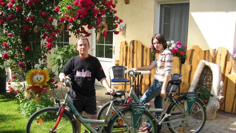 Two people with bicycles in front of a house with flowers and wooden decorations.