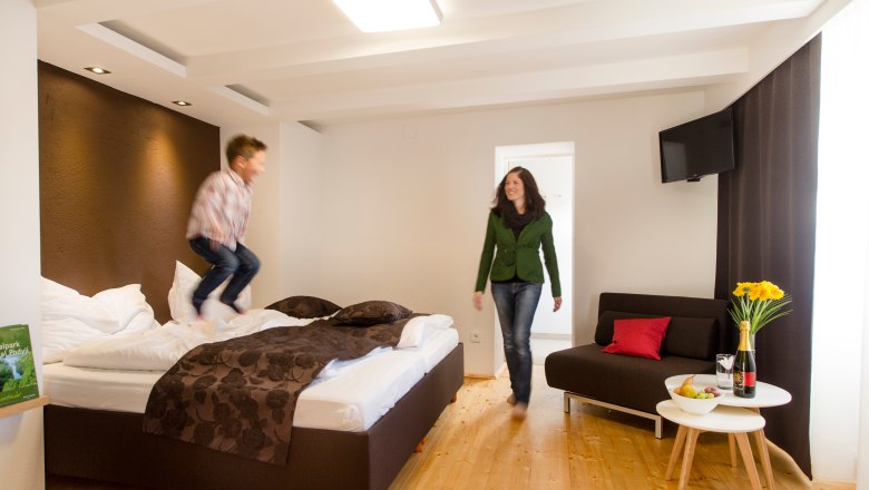 A child jumps on a bed in a modern hotel room while a woman looks on smiling. A table with flowers and wine stands next to it.