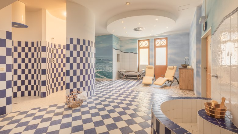 Interior view of a wellness area with blue and white tiles, deckchairs and a wooden bucket.
