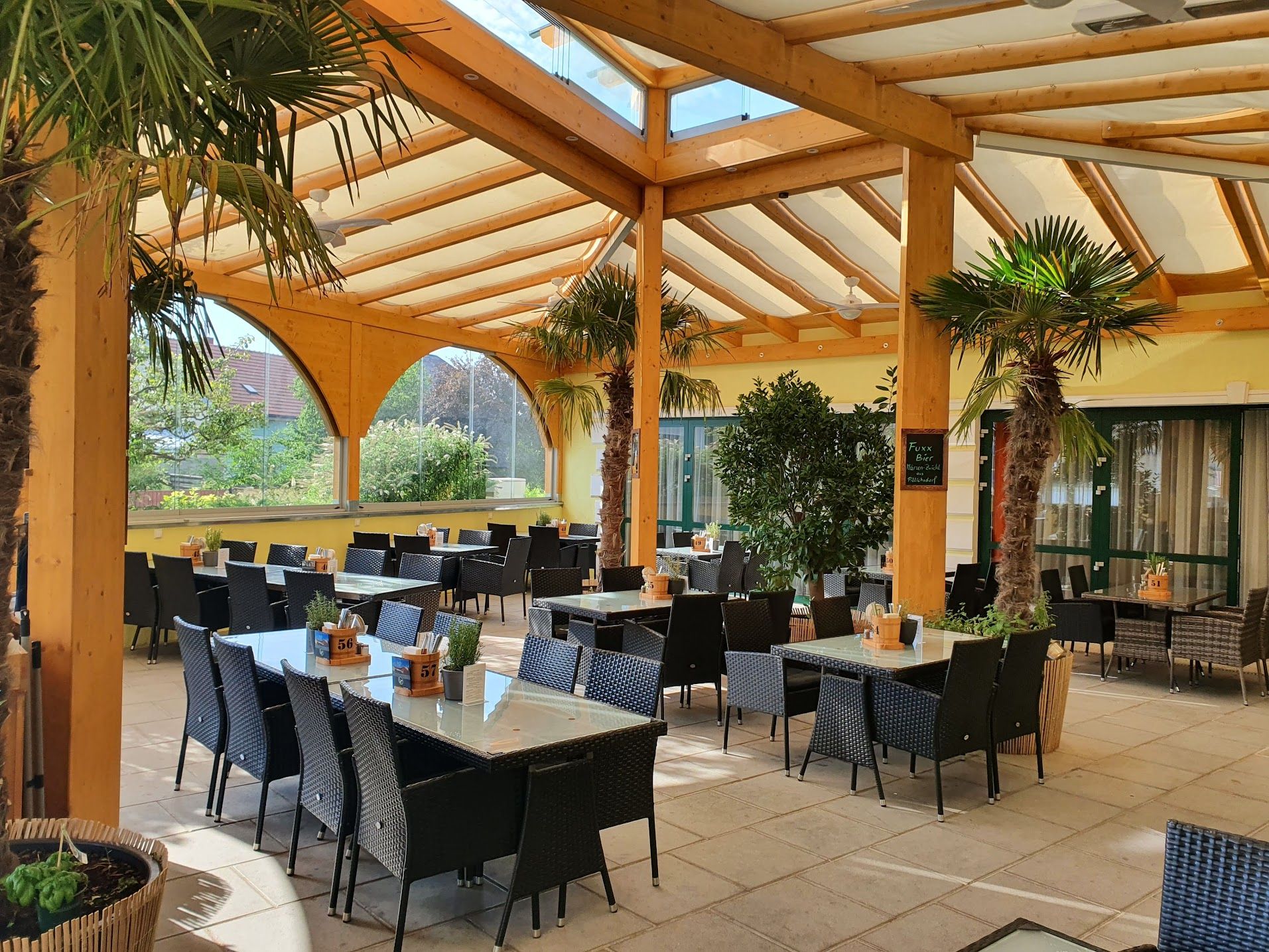 Covered terrace of a restaurant with palm trees and tables.