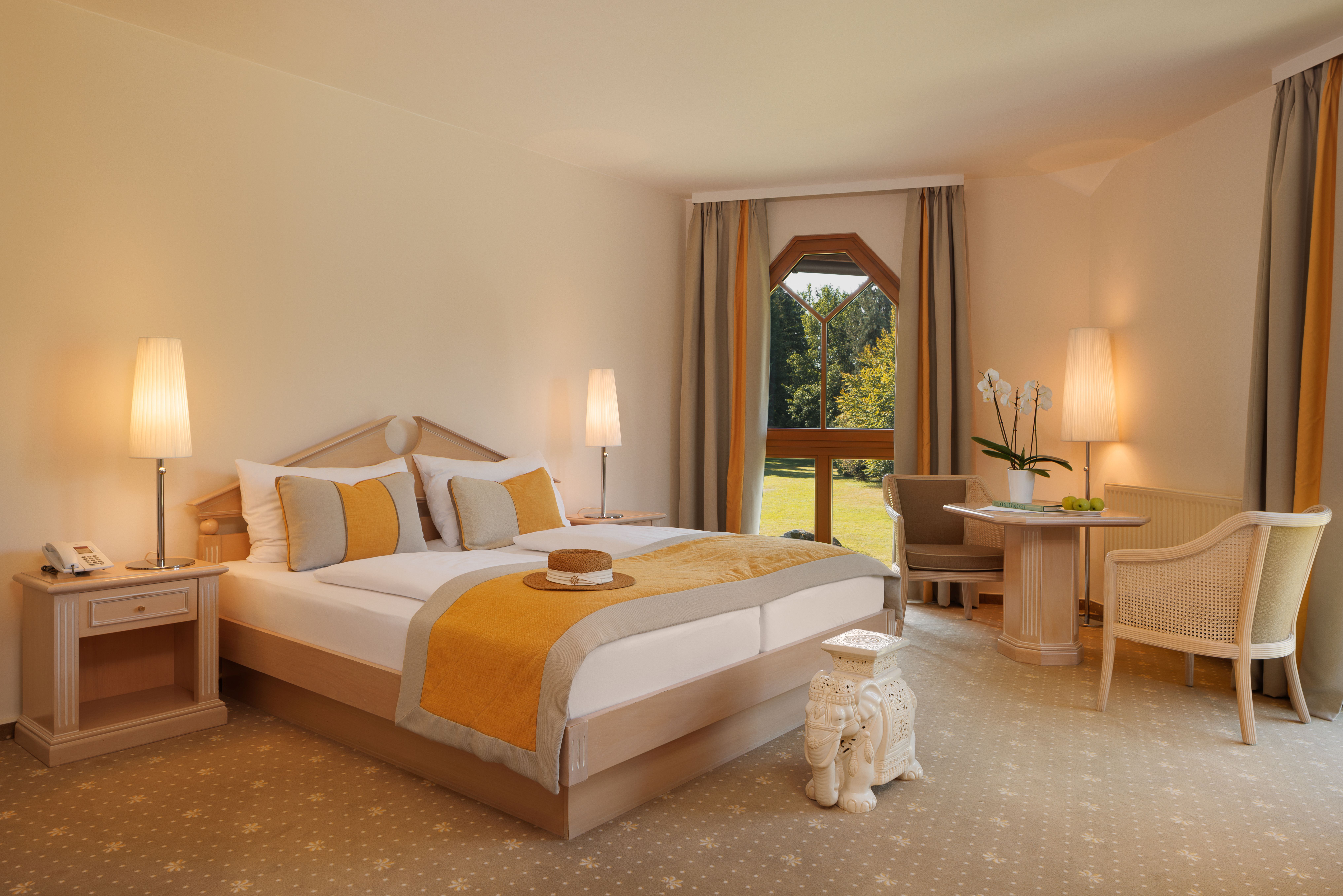 Elegant hotel room with double bed, yellow accents and large windows overlooking a garden.