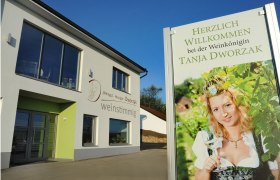 A modern building with a sign showing a woman in traditional costume with a wine glass. Text: Welcome to the wine queen Tanja Dworzak.