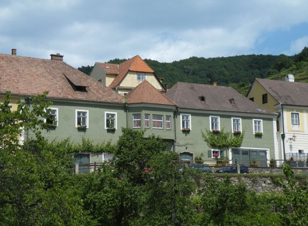 Terraced houses with green facades and red roofs in front of wooded hills.