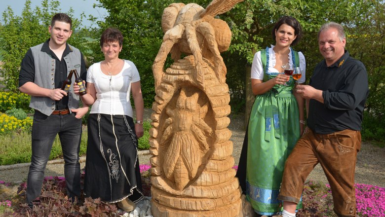 Four people in traditional dress stand next to a wooden sculpture outdoors.