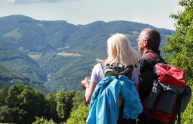 Two hikers with rucksacks look out over a mountainous landscape.