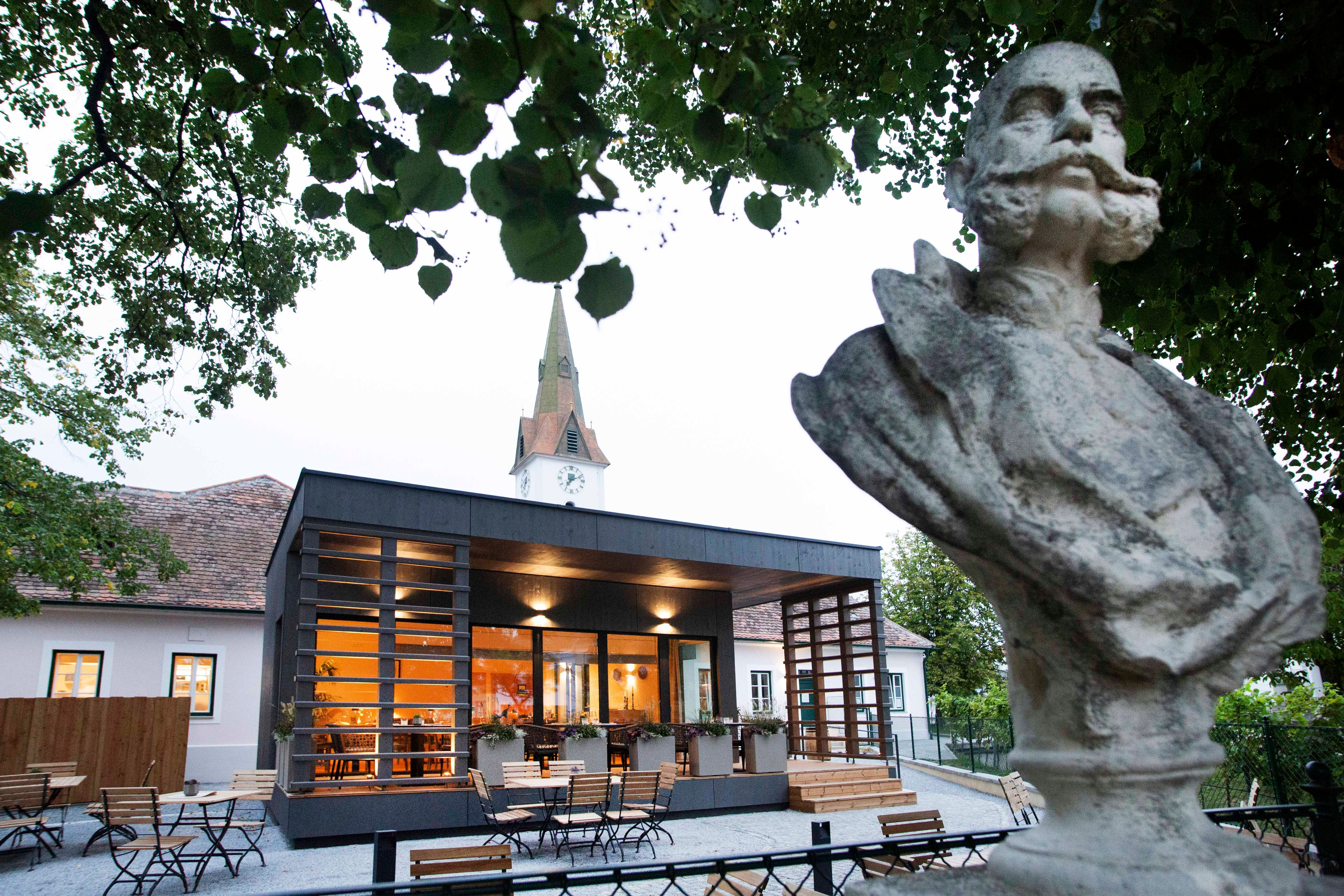 Bust in front of a modern building with a church tower in the background, surrounded by trees.
