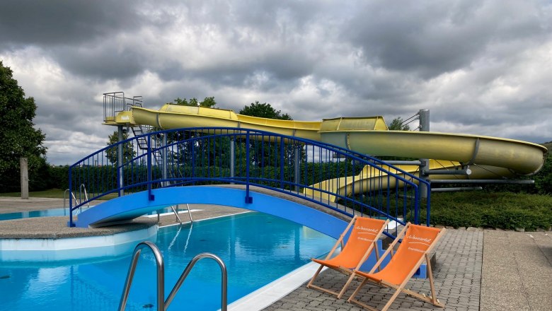 Two orange deckchairs stand next to a swimming pool with a blue bridge and a yellow water slide in the background.
