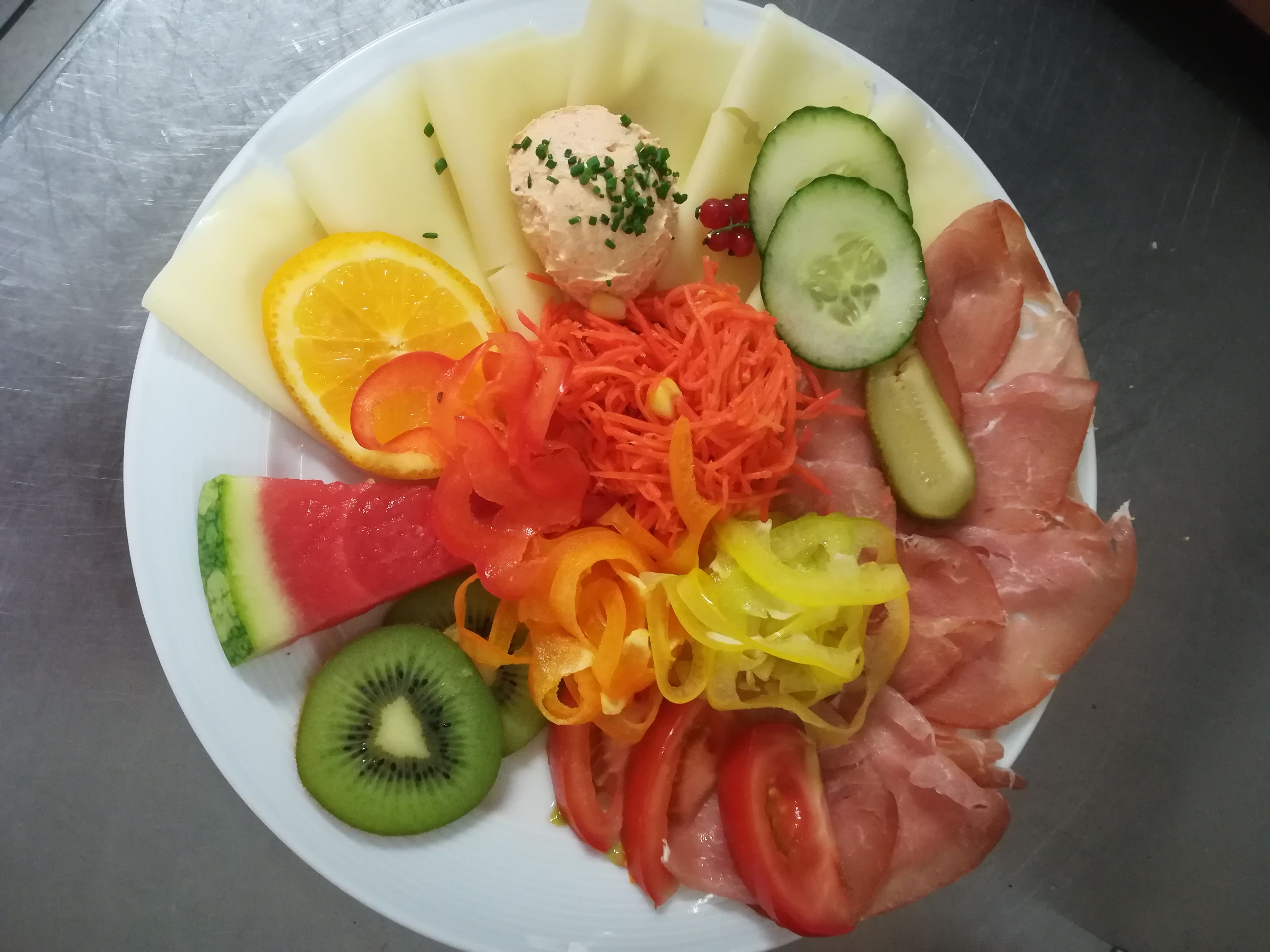 A plate with cheese, ham, vegetables and fruit, including melon, kiwi, cucumber and peppers.