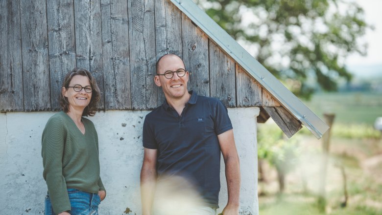 Two people stand smiling in front of a wooden wall outside.