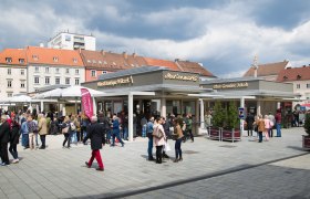 Crowd on a busy marketplace with stalls and buildings in the background.