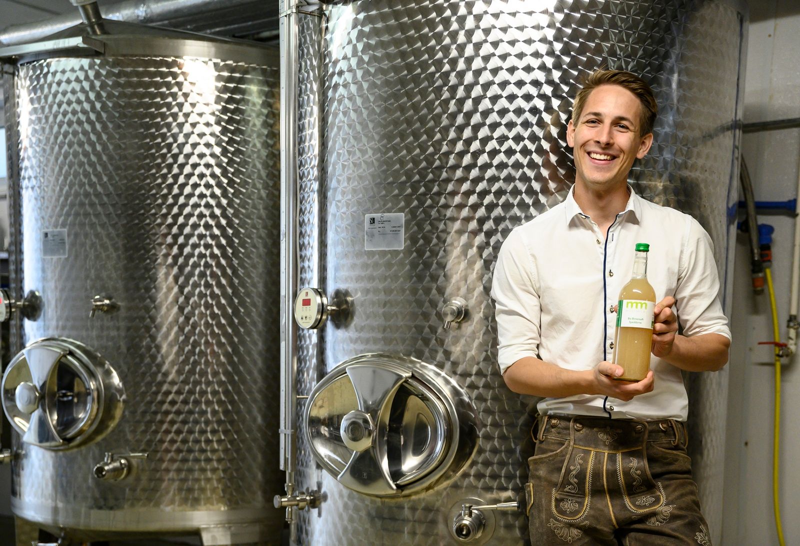 Man in traditional clothing holding a bottle in front of large metal tanks.