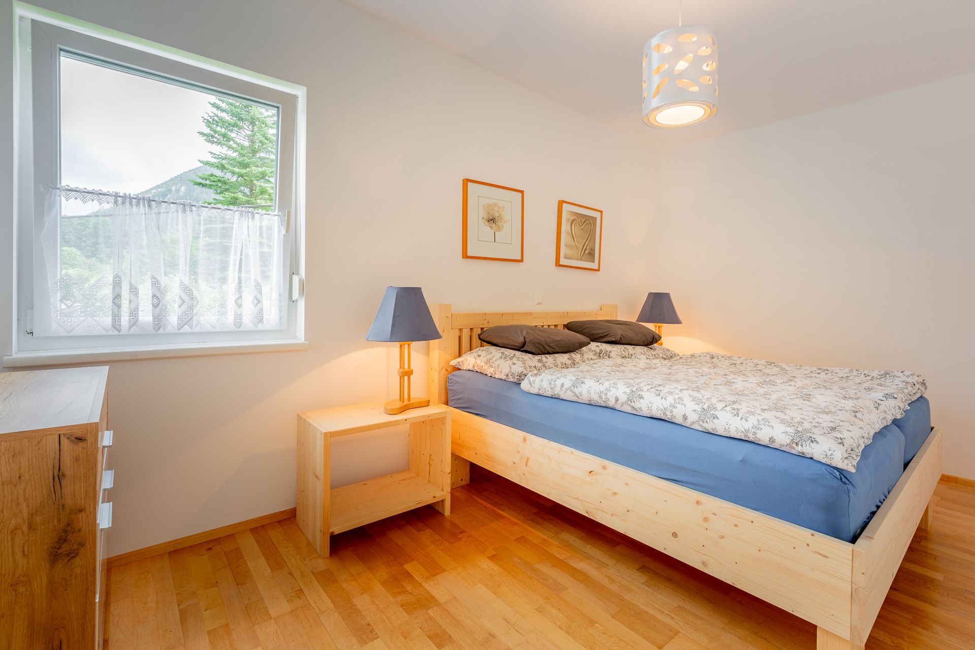 A cozy bedroom with a wooden bed, blue bed linen and two bedside lamps. A window with a view of trees and mountains.