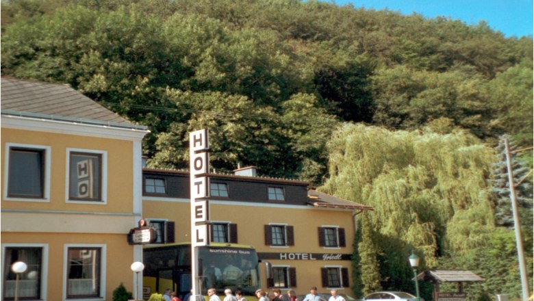 House, © Hotel-Gasthof Friedl A yellow hotel building with a large hotel sign and people in front of it, surrounded by trees.
