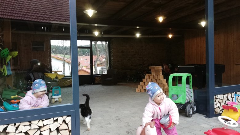 A child in winter clothes plays with a toy horse in an open room with piles of wood and toys. A cat walks past.