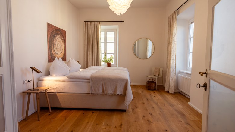 Cozy bedroom with double bed, wooden floor, wall art, round mirror and plants on the windowsill.