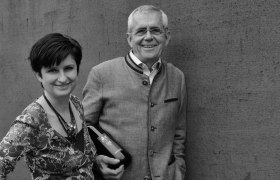 A man and a woman stand smiling in front of a wall. The man is holding a bottle of wine.