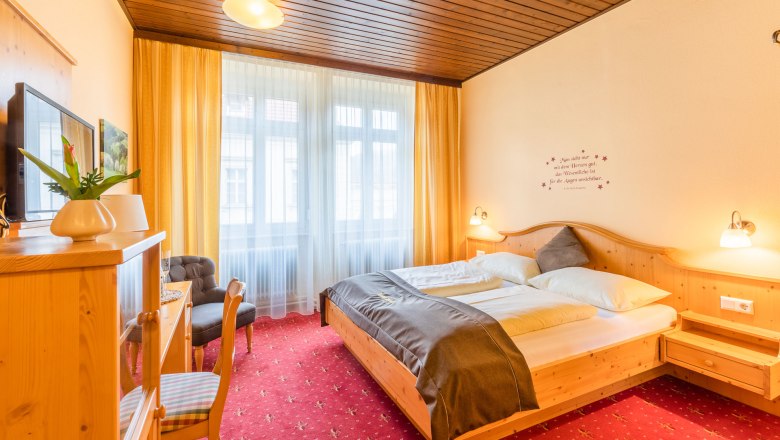 Comfortable hotel room with double bed, wooden furniture and red carpet.