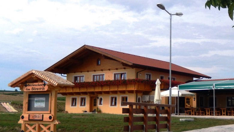 A traditional inn with a wooden balcony and floral decorations, next to a sign reading 'Buschenschank Fam. Böswirth'.