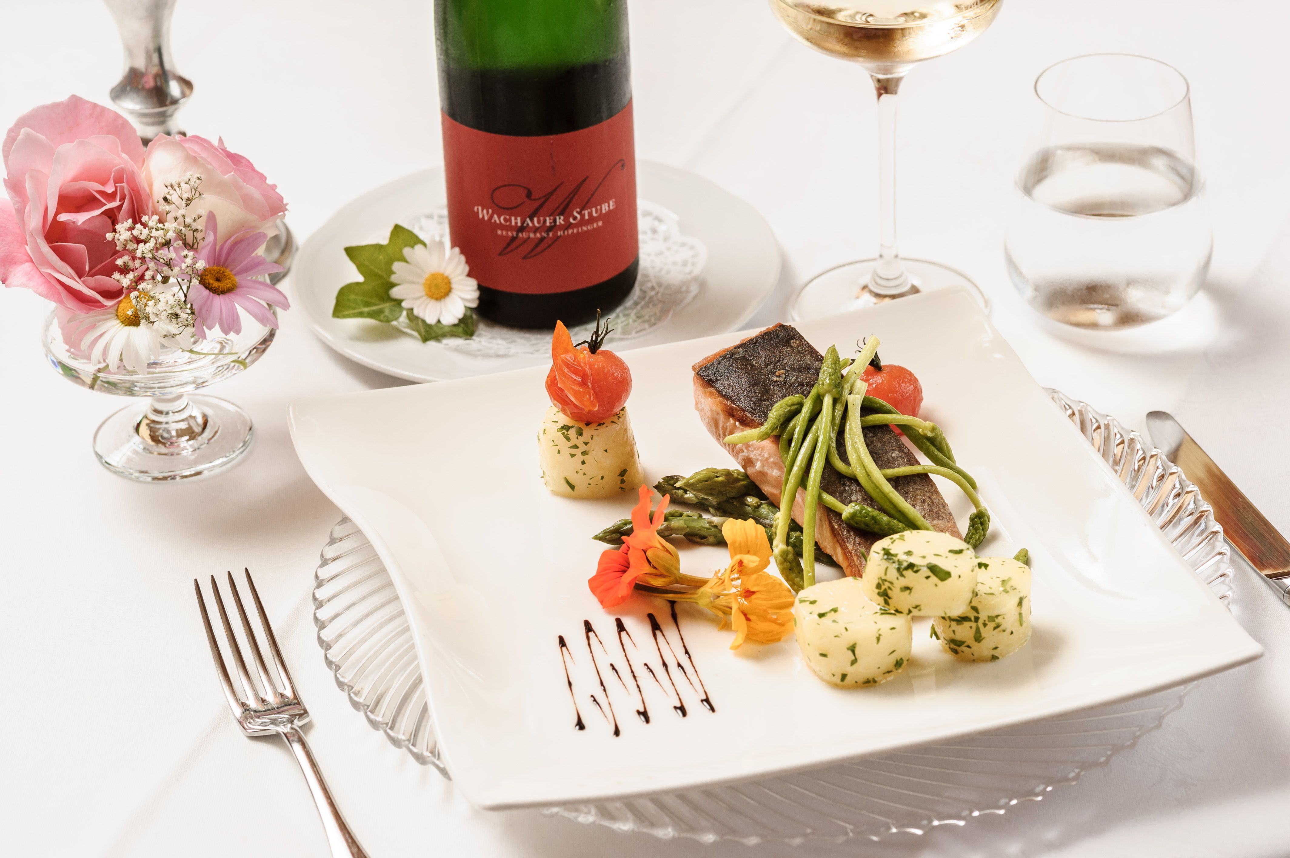 Elegant dish with fish, asparagus and potatoes on a white plate, decorated with flowers. A bottle of wine and a glass in the background.
