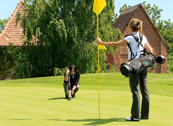 Two women playing golf on a green golf course, one holding the flag, the other aiming at the hole.