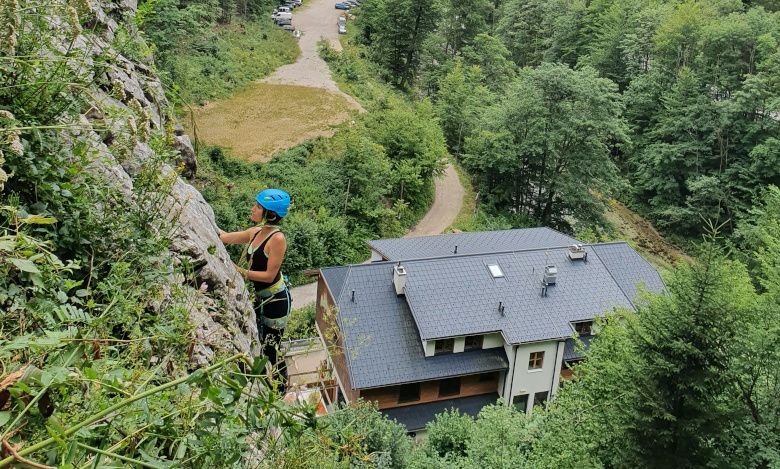 Person climbing on a rock face with helmet and climbing equipment, buildings and forest in the background.