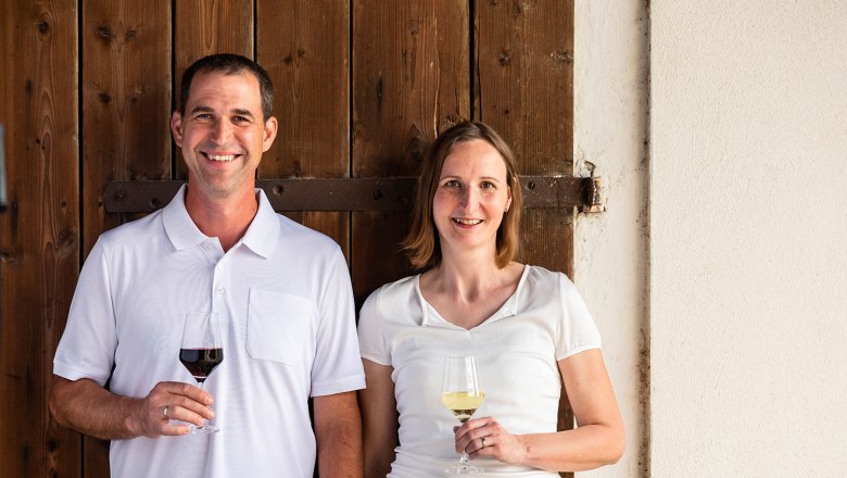 A man and a woman stand smiling in front of a wooden wall, both holding glasses of wine.