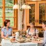 Three women sit at a laid breakfast table in a cozy room with wooden furniture and large windows overlooking vineyards.