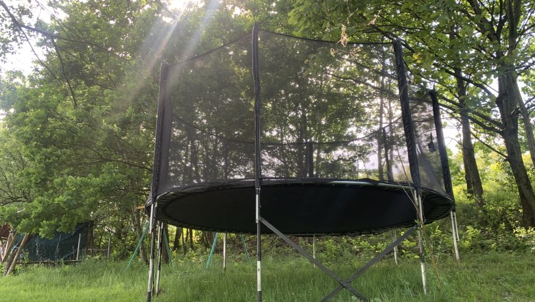 A trampoline with a safety net stands in a meadow surrounded by trees. Rays of sunshine shine through the canopy.
