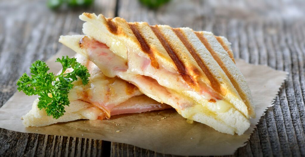 A grilled sandwich with ham and melted cheese, garnished with parsley, on a wooden board.