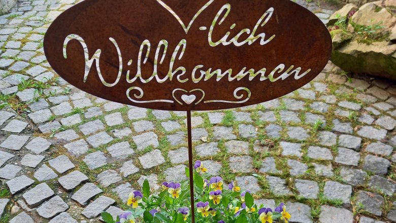 Metal sign with 'Welcome' above a pot of yellow-purple flowers on a cobbled path.