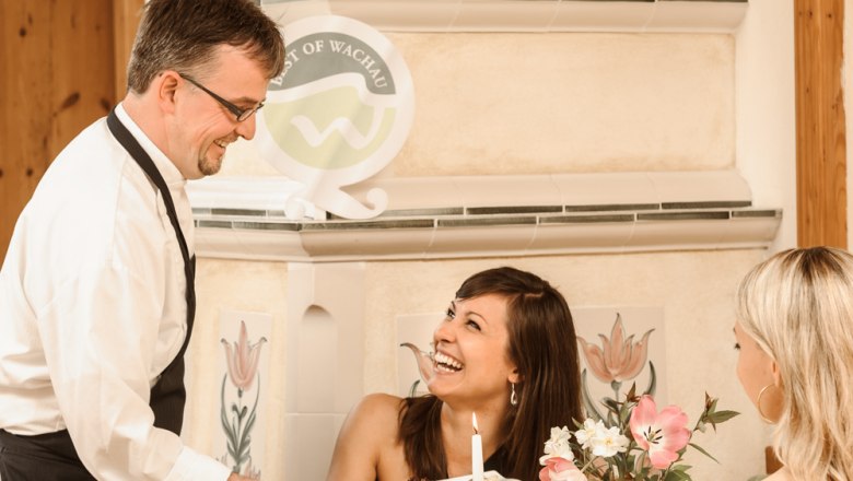 A waiter serves two women in a cozy restaurant. One woman laughs as she accepts the food. The table is elegantly set.