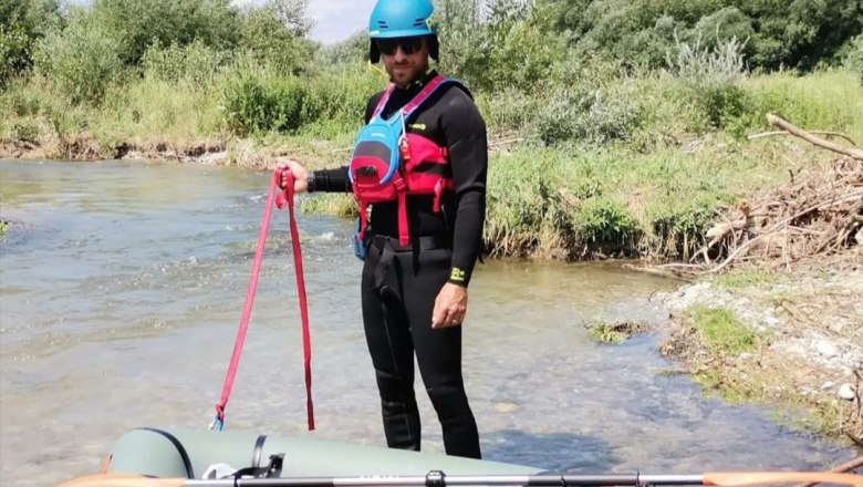 A person in a wetsuit and helmet stands next to an inflatable boat on a riverbank.