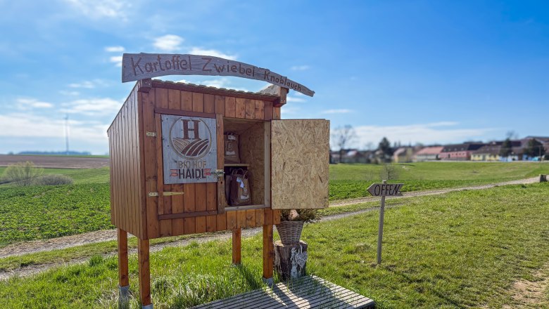 A small stall on a country lane with the inscription 'Kartoffel Zwiebel Knoblauch' and 'Bio Hof Haide'. A sign says 'Open'.