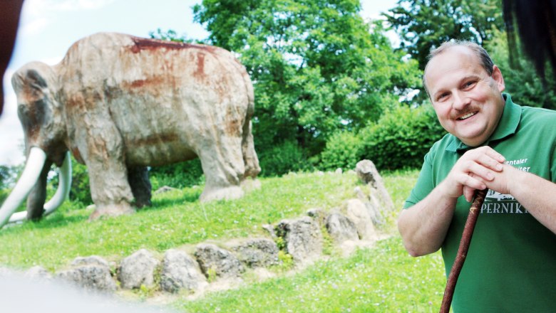 A man smiles in front of a large mammoth statue outdoors.