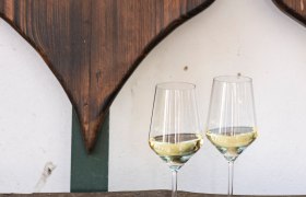 Two wine glasses with white wine on a wooden table in front of a wooden wall with heart decoration.