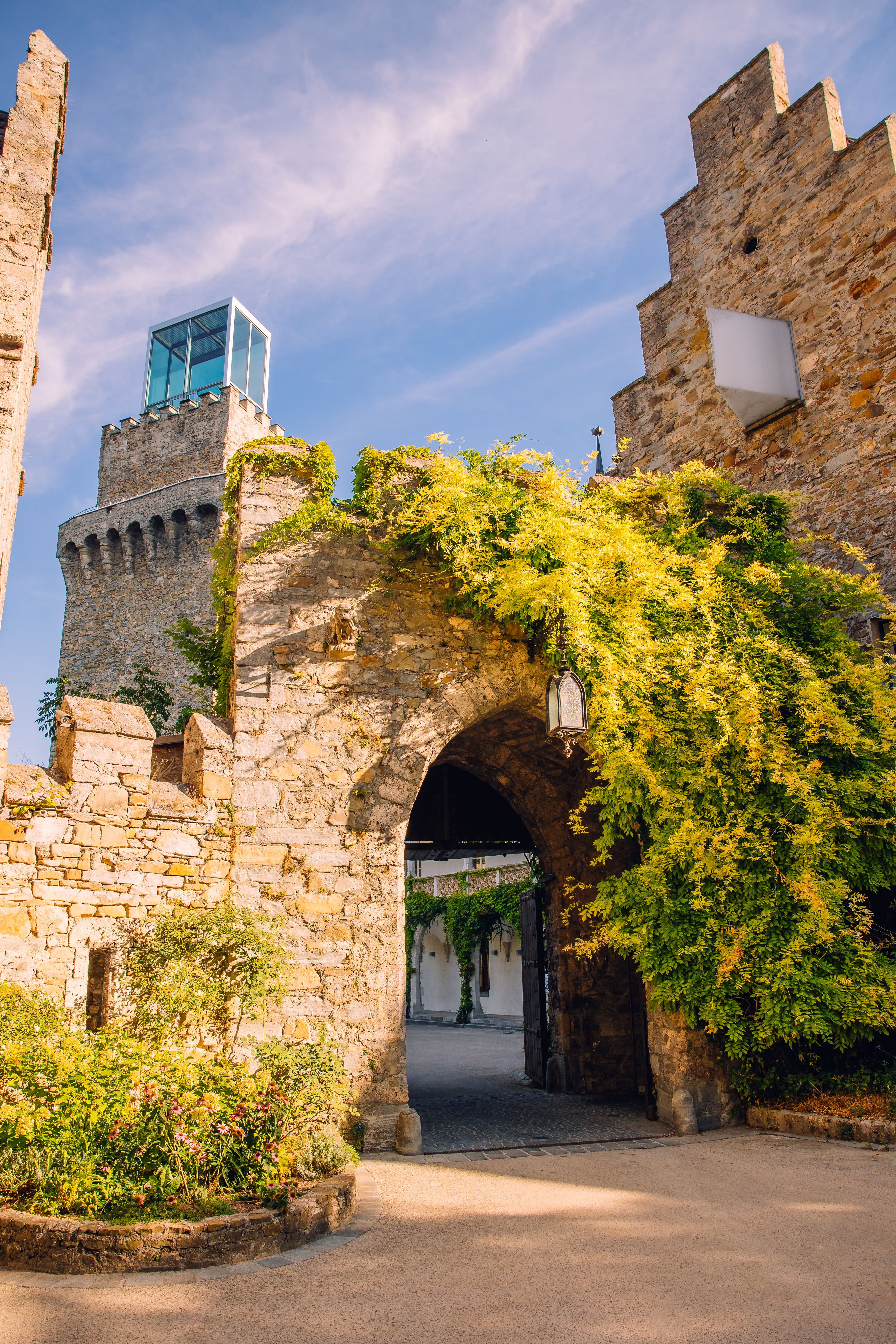 The historic archway of Schloss Rothschild in Waidhofen an der Ybbs, featuring stone walls covered in greenery and a modern glass structure atop the castle tower.