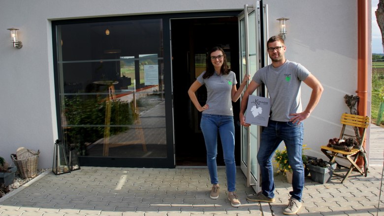 Two people are standing in front of a building with a glass door, both wearing gray T-shirts and jeans. One person is holding a sign saying 'Kornherr'.
