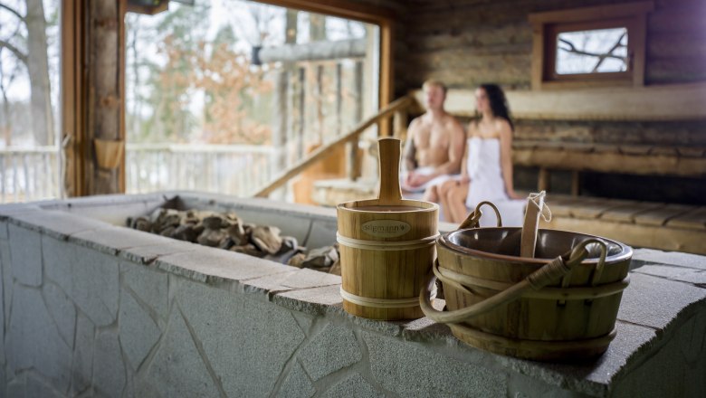 Interior view of a sauna with two people sitting on wooden benches in the background. Sauna buckets and stones can be seen in the foreground.