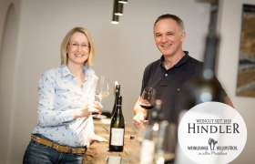 A pair of winemakers stand smiling with wine glasses in their hands at a table in the Hindler winery.