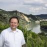 Man standing in a vineyard with river and hills in the background.
